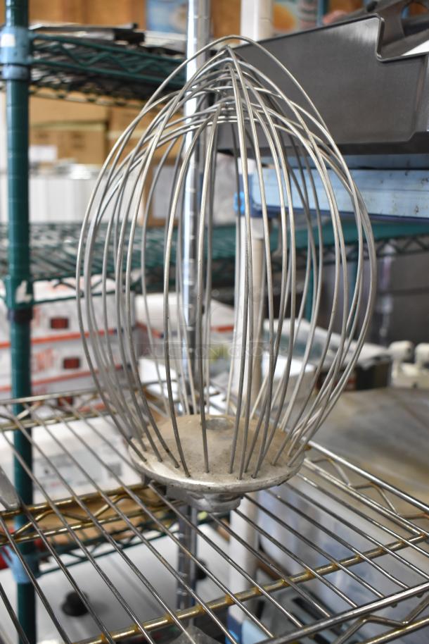 Hobart mixer whisk attachment, metal construction, visibly used condition, set on a metal shelf.