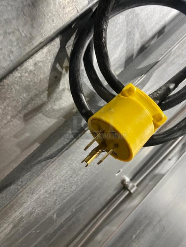Yellow electrical plug with worn prongs attached to a black cable, resting against a textured metal surface.