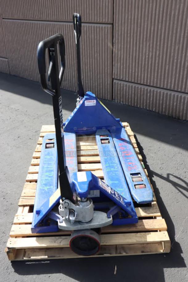 Pair of Lavex pallet jacks in blue, shown on a wooden pallet. Handles upright, visible wear, sturdy construction.
