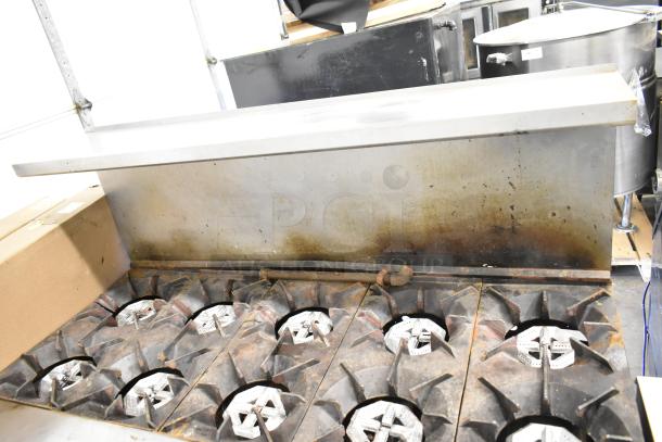 Commercial gas-powered 10 burner range with two ovens, overhead shelf, and backsplash. Used condition, visible wear.