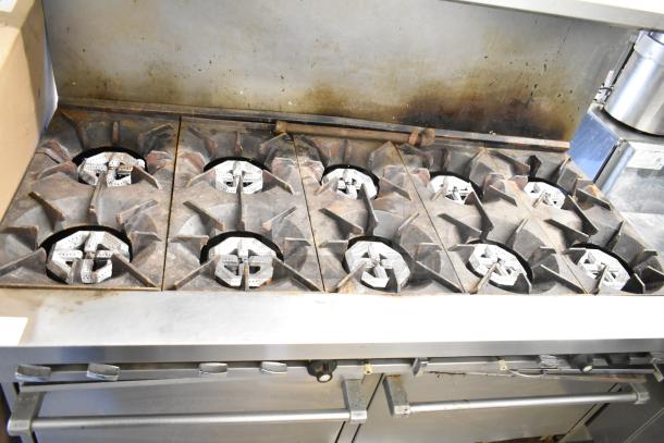 Commercial gas-powered 10-burner range with 2 ovens, overhead shelf, and backsplash. Some visible wear. Ideal for professional kitchens.