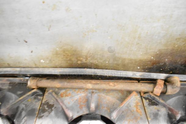 Rusty component of commercial gas range, highlighting wear. Part of 10-burner range with ovens, shelf, and backsplash.