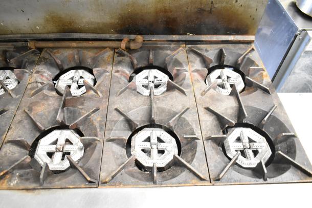 Commercial gas-powered 10-burner range with two ovens, shelf, and backsplash. Features visible wear, ideal for heavy-duty cooking.