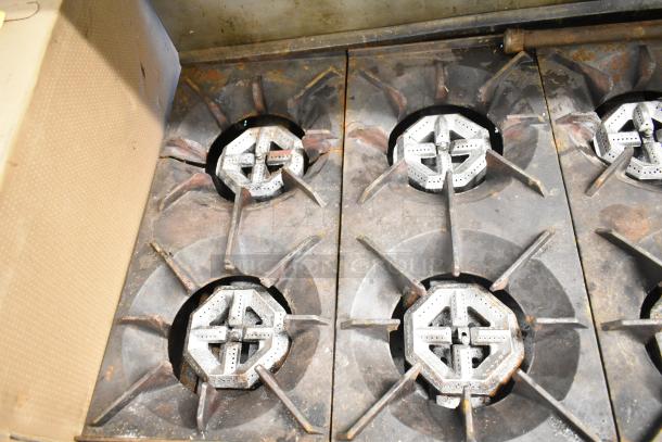 Commercial gas-powered 10 burner range with 2 ovens, over shelf, and backsplash. Shows signs of wear, suitable for heavy-duty use.