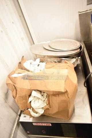 Nine assorted items including stacked metal pizza pans and lightbulbs in a damaged cardboard box on a Blodgett appliance.