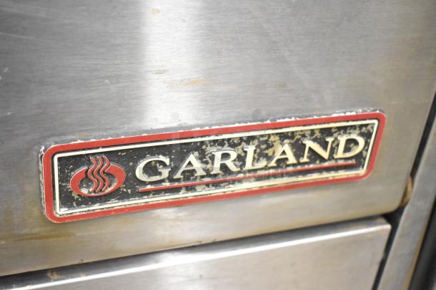 Garland commercial gas-powered range, 6 burners, flat top griddle, 2 ovens, over shelf, back splash, visible logo.