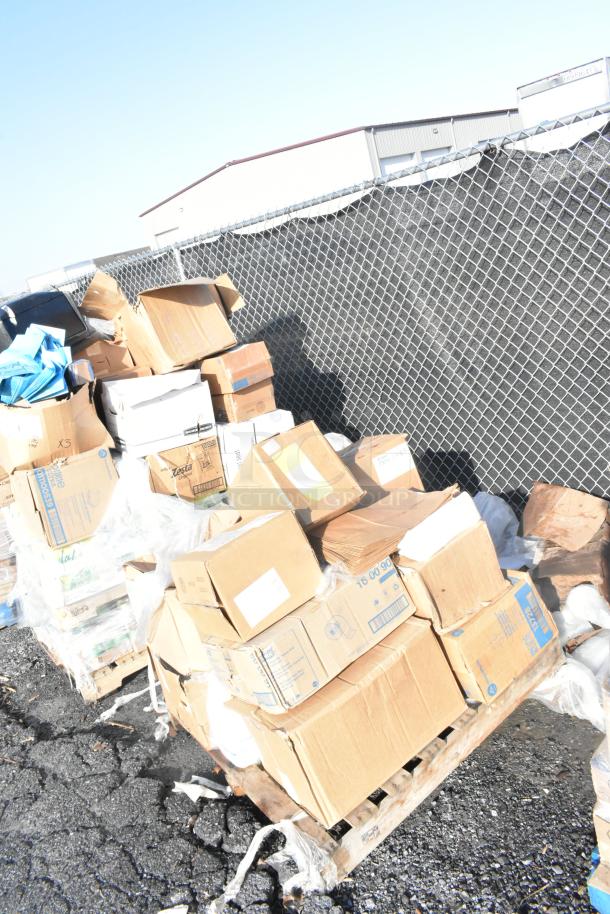 Pile of cardboard boxes on a pallet, labeled with various shipping labels, positioned outdoors near a metal fence.