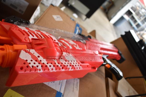 Red and orange toy gun with a textured design, marked "MXVI-10K," placed on cardboard boxes. Additional items not visible.
