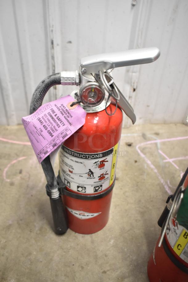 Red fire extinguisher with silver nozzle, attached hose, and inspection tag indicating service details.