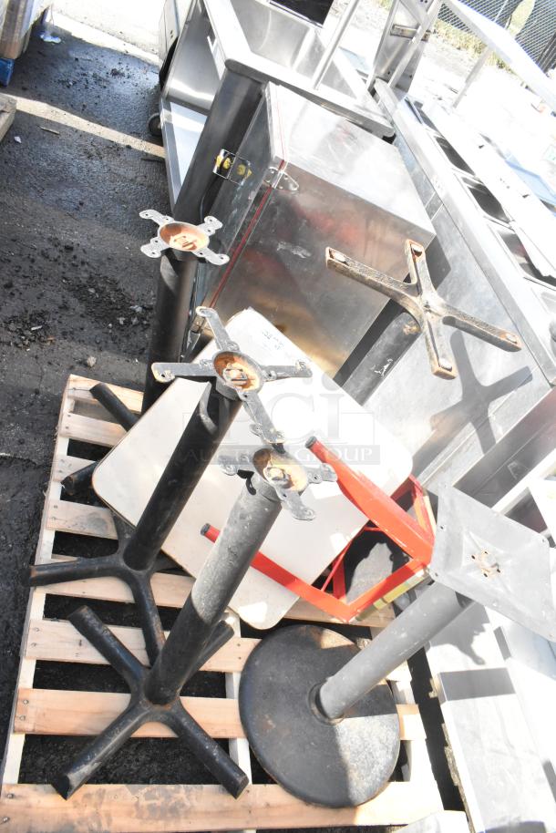 Five black metal table bases with cross-shaped tops on a wooden pallet. Used condition, rust visible.