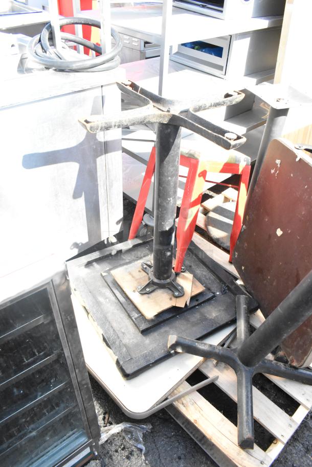 Five metal table bases with square and round tops, stored on a pallet. Tables show signs of wear.