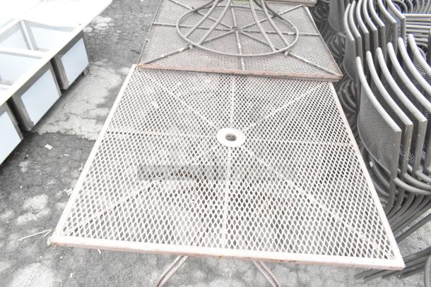 16 metal mesh patio tables with umbrella holes, stacked, in used condition, on asphalt surface.