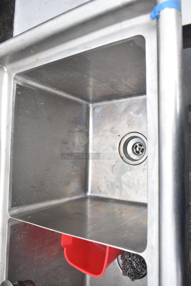 Commercial 3 bay stainless steel sink, used condition. Visible drain, red plastic cup, cleaning scrubber in sink basin.