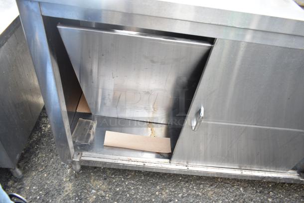 Commercial stainless steel prep station with under shelves, featuring sliding doors and slight wear. Suitable for kitchen use.