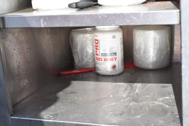 Stainless steel commercial table with under shelves, showing storage space with a whey protein container and wrapped items. Slightly worn condition.