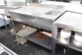 Commercial stainless steel table with sink bay and under shelf, showing signs of use; sturdy design for kitchen settings.