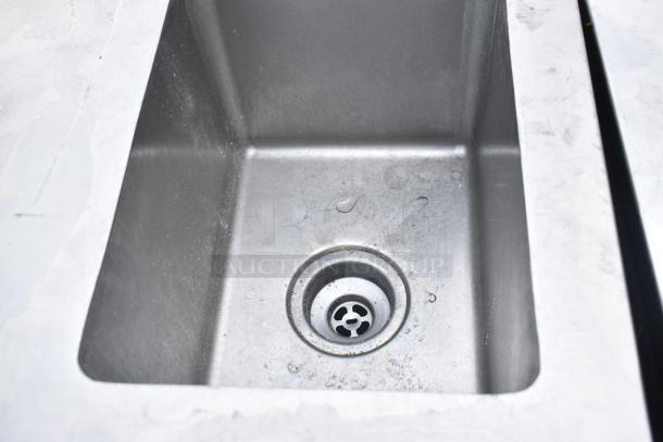 Metal sink bay within a commercial table, featuring visible drain. Some surface wear present. Ideal for kitchen use.