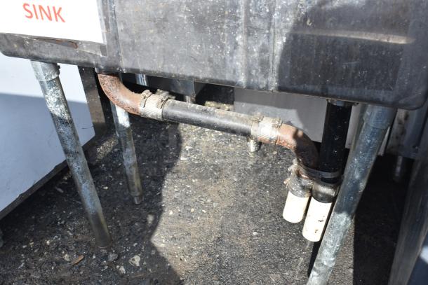 Commercial 3 bay sink with metal legs and visible plumbing connections, showing signs of wear. Label indicates "SINK."