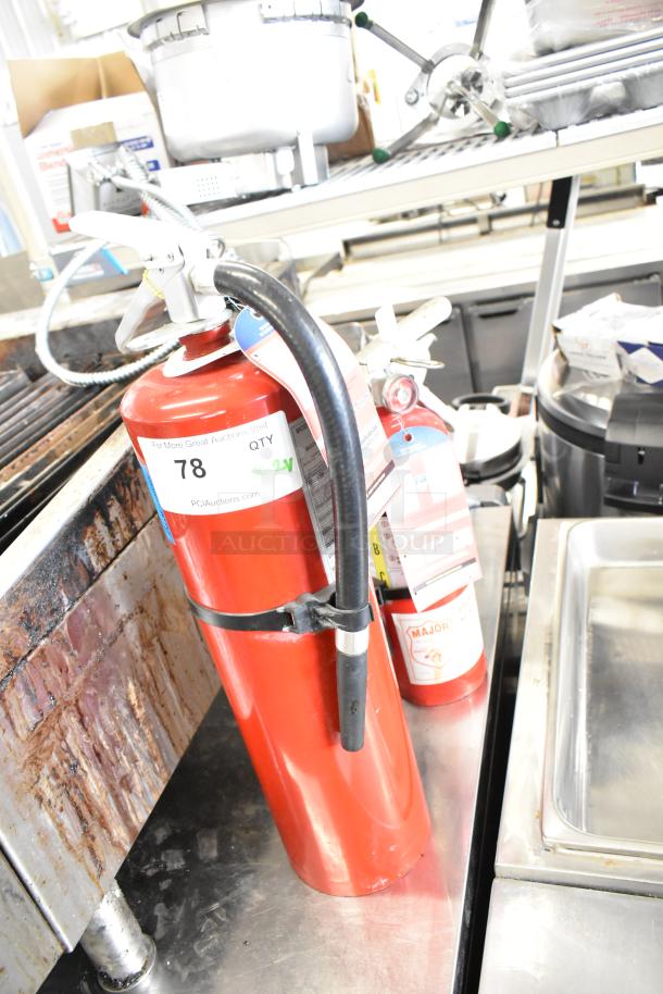 Two red fire extinguishers, including Buckeye brand, on a metal counter. Labels indicate auction lot number 78.