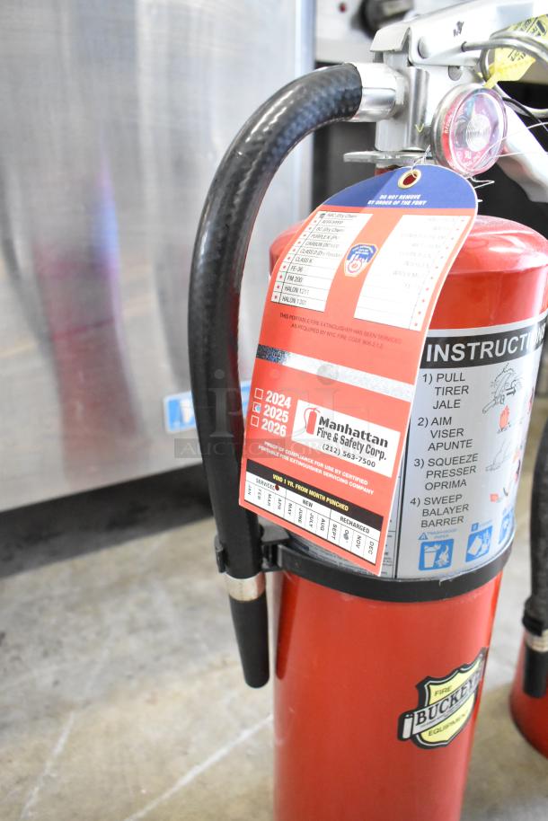 Two red fire extinguishers, brand Buckeye, inspection tags from Manhattan Fire & Safety, not available for shipping.