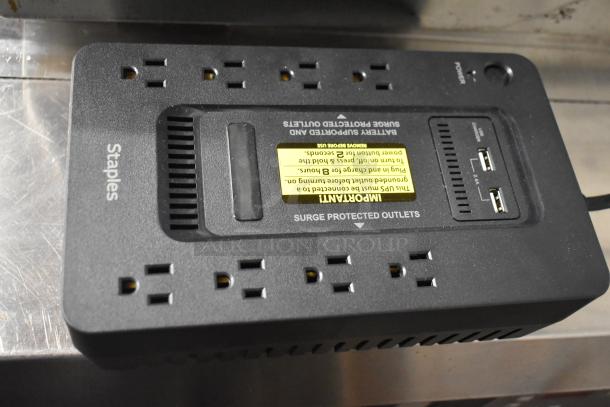 Staples uninterruptible power supply with multiple surge-protected outlets and USB ports, black, visible yellow warning label.