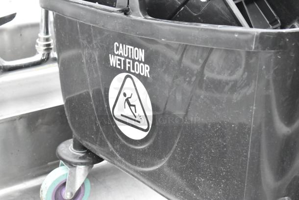 Black poly mop bucket with wringing attachment, features "Caution Wet Floor" label, sturdy wheels. Slightly used condition.