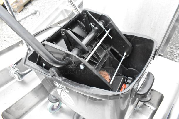 Black poly mop bucket with wringing attachment, caution wet floor label, durable, with wheels for easy mobility.