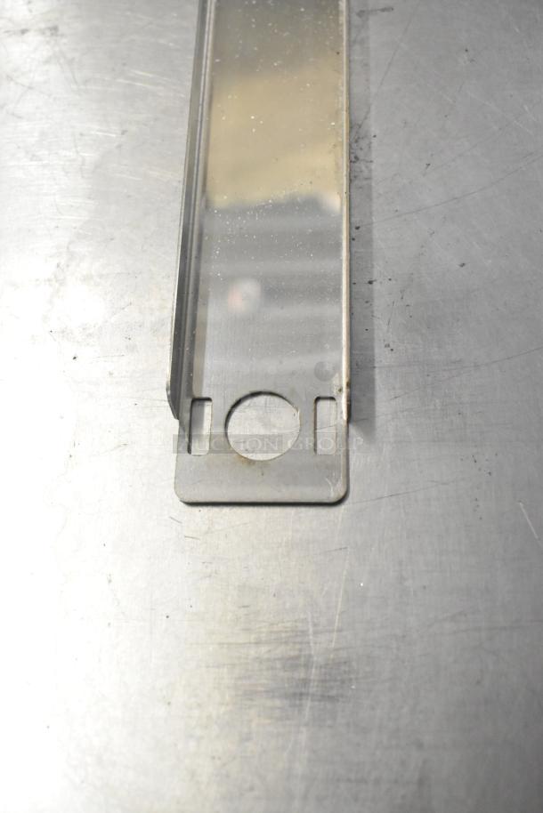 Silver metal component, possibly a part of Cadco/Unox equipment, featuring a slotted design with circular cutout, on a brushed metal surface.