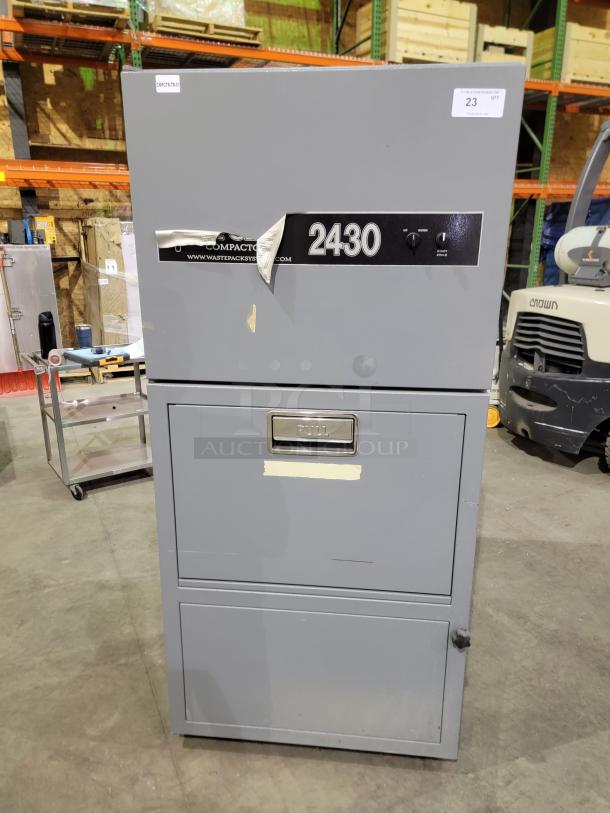 FOX 2430 industrial waste compactor in grey, featuring two-dial controls, steel frame, and bin on casters. Model label visible.