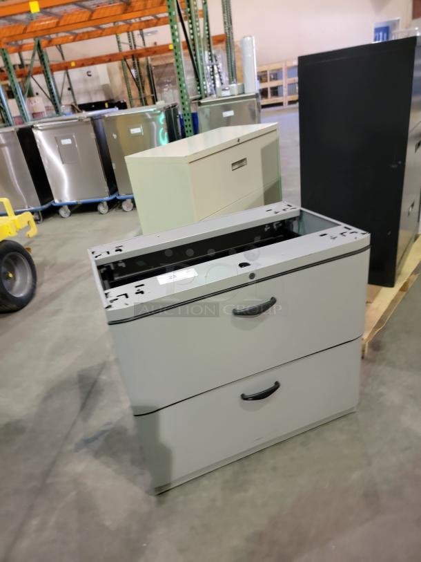 Mixed lot of three steel lateral file cabinets, featuring a black two-drawer, a white three-drawer, and another black two-drawer unit, all with smooth drawer glides, in a warehouse setting.
