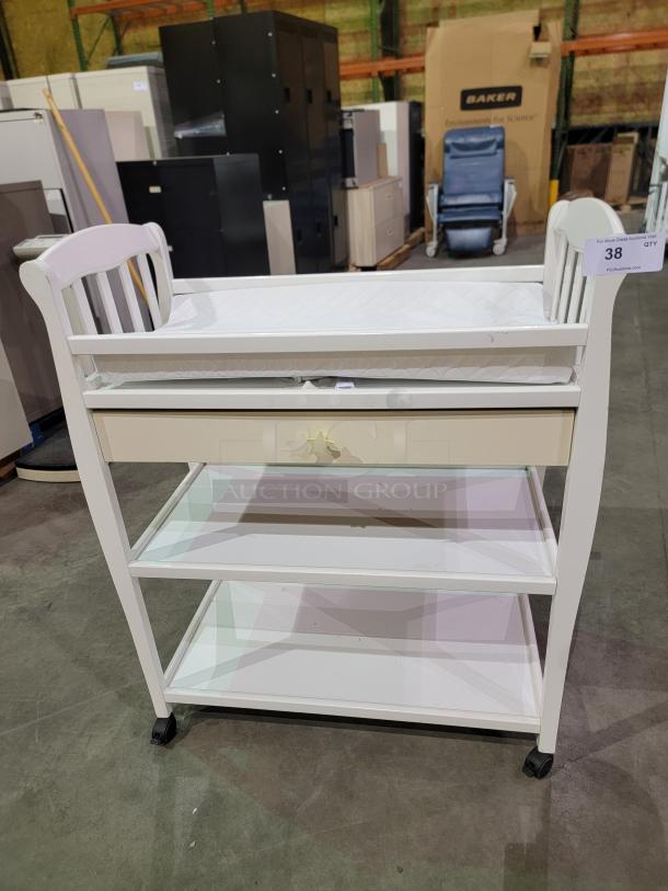 White Sorelle Manhattan changing table with padded top, star knob drawer, two shelves, and casters for easy mobility.