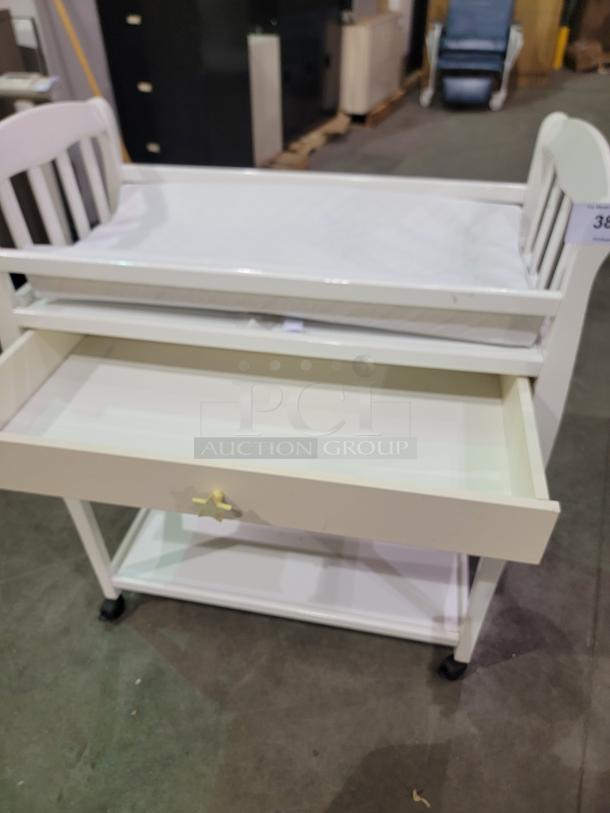 White Sorelle Manhattan changing table with padded mat, star-shaped drawer knob, two open shelves, and casters for mobility.