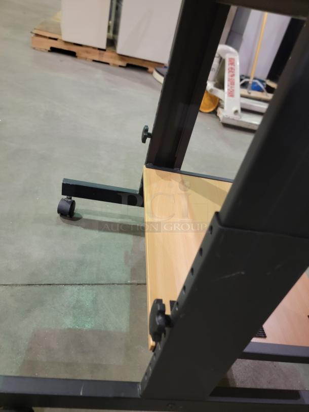 Mobile computer cart in a warehouse, featuring a black metal frame, lower wooden shelf, and caster wheels for mobility.