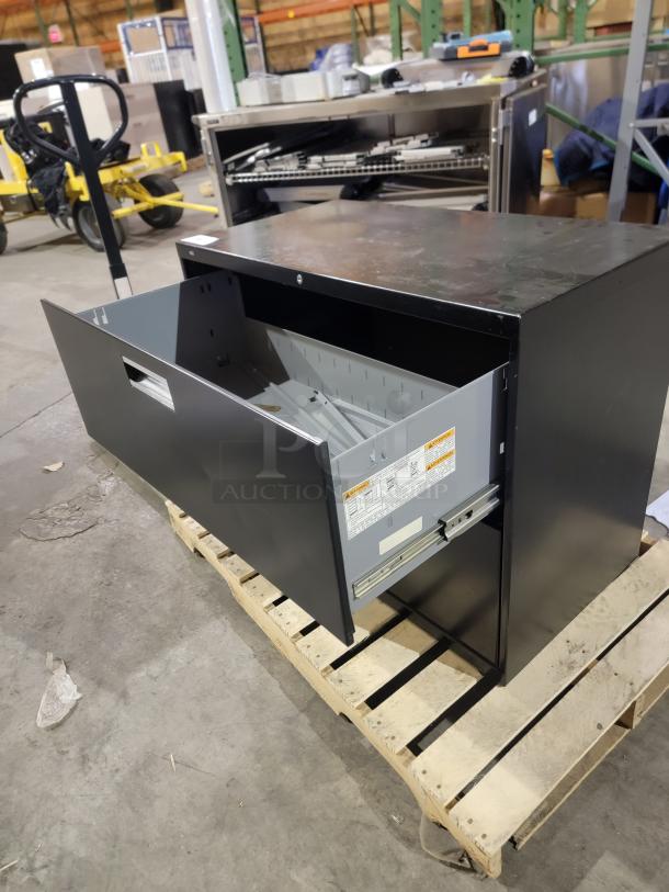 HON Brigade 600 Series 2-drawer lateral file, black finish, open drawer on pallet, visible safety labels.