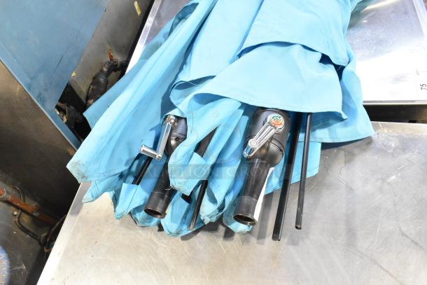 Two blue patio umbrellas with crank handles, folded, and placed on metal surface. Auction item, bid includes both.
