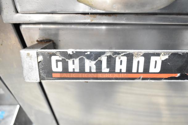 Garland commercial gas range, 4 burners, oven, overshelf, backsplash. Label shows wear. Stainless steel exterior.