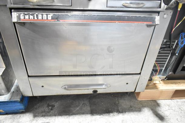 Garland commercial gas-powered 4-burner range with oven, visible oven door with brand logo. Features shelf and backsplash.