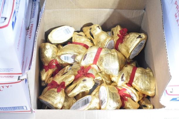 Box of Lindt Gold Bunny chocolates, wrapped in gold foil with red ribbons, stacked in a cardboard box. Auction lot includes 10 boxes.