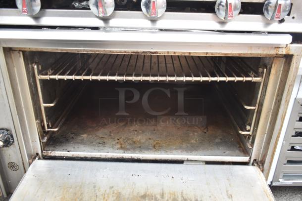 Commercial gas-powered 12 burner range with open oven, showing interior; includes over shelf, back splash, and casters.