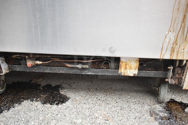 Commercial gas-powered 12 burner range with 2 ovens, over shelf, and backsplash on casters. Visible wear and rust.