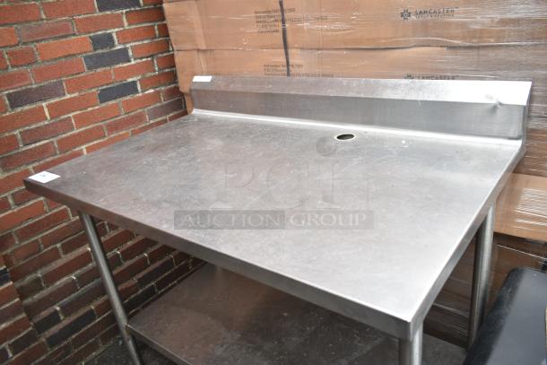Stainless steel table with backsplash and under shelf, features hole on top surface, in good condition, industrial setting.