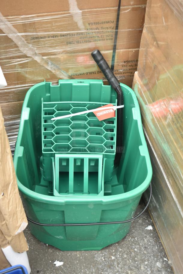 Green Sparta mop bucket with black handle, scratch and dent label, new condition, placed among cardboard boxes.