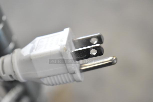 Close-up of a white electrical plug with two metal prongs, likely part of store display units for auction, featuring Soundcore and Sony speakers.