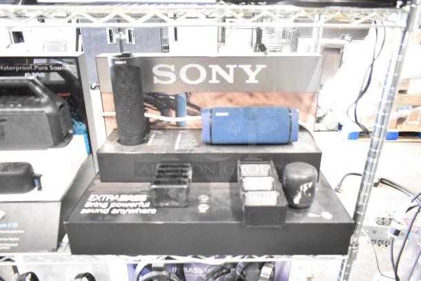 Store displays featuring a Soundcore speaker and Sony speakers in black and blue. Labels highlight powerful sound features. Condition: Displayed but good.