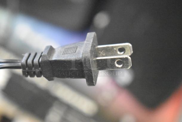 Close-up of a black power plug. Background shows blurred electronic devices. Part of store display auction item set.