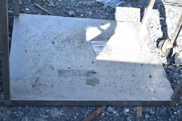 Commercial table with metal under shelf, showing signs of wear with dirt and shadow on surface, set on rocky ground.