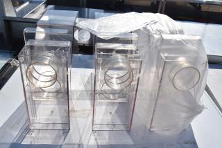 Three clear acrylic hoppers with circular metal rings, used condition, set on a white surface. Suitable for bid.