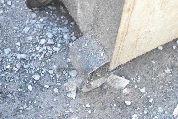 A close-up of a wooden crate corner with a metal bracket on a gravel surface. Condition detail for auction.