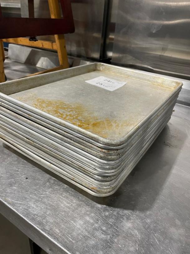 Stack of commercial aluminum baking sheet pans, used condition, visible wear marks, auction item tag attached.
