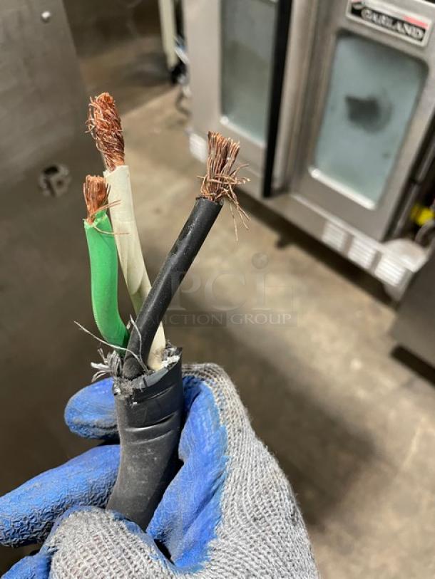 Three exposed electrical wires in a gloved hand, near a commercial kitchen appliance.